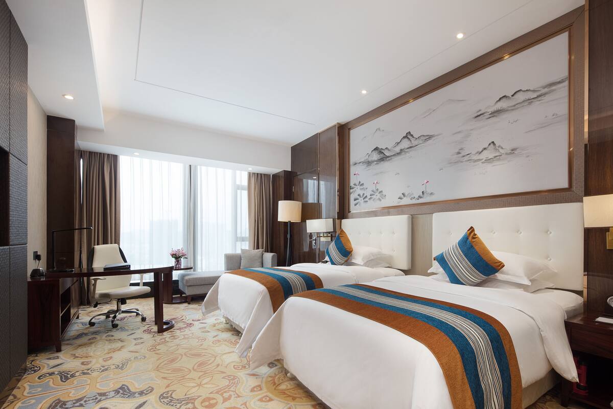 Quarto de hotel com duas camas, uma escrivaninha, uma cadeira, um abajur e um quadro grande na parede.