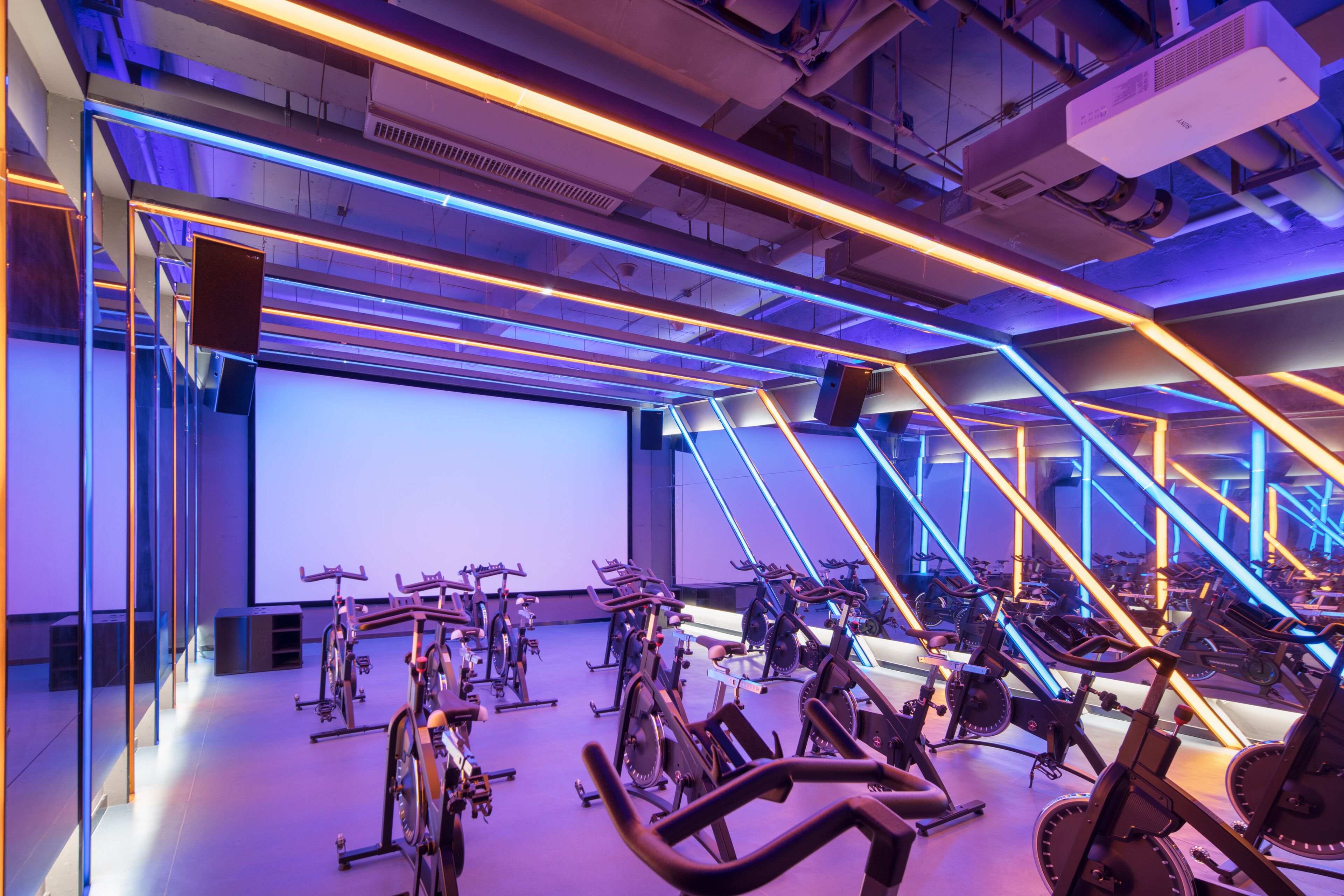 A modern fitness center with multiple stationary bikes arranged in rows, a large screen, and a ceiling with exposed ductwork and lighting.