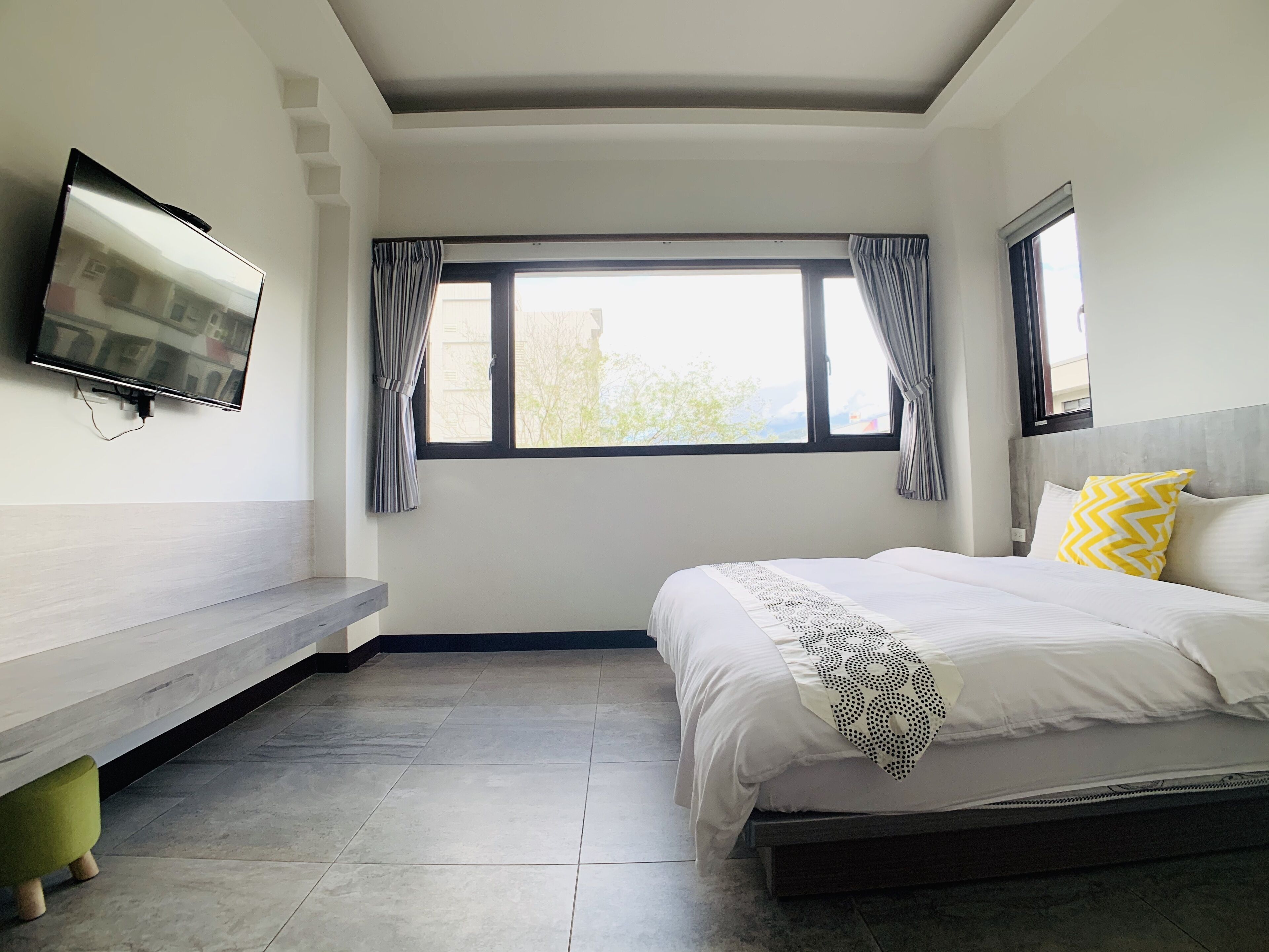 A modern hotel room with a bed, a flat-screen TV mounted on the wall, a window with curtains, and a bench by the window.