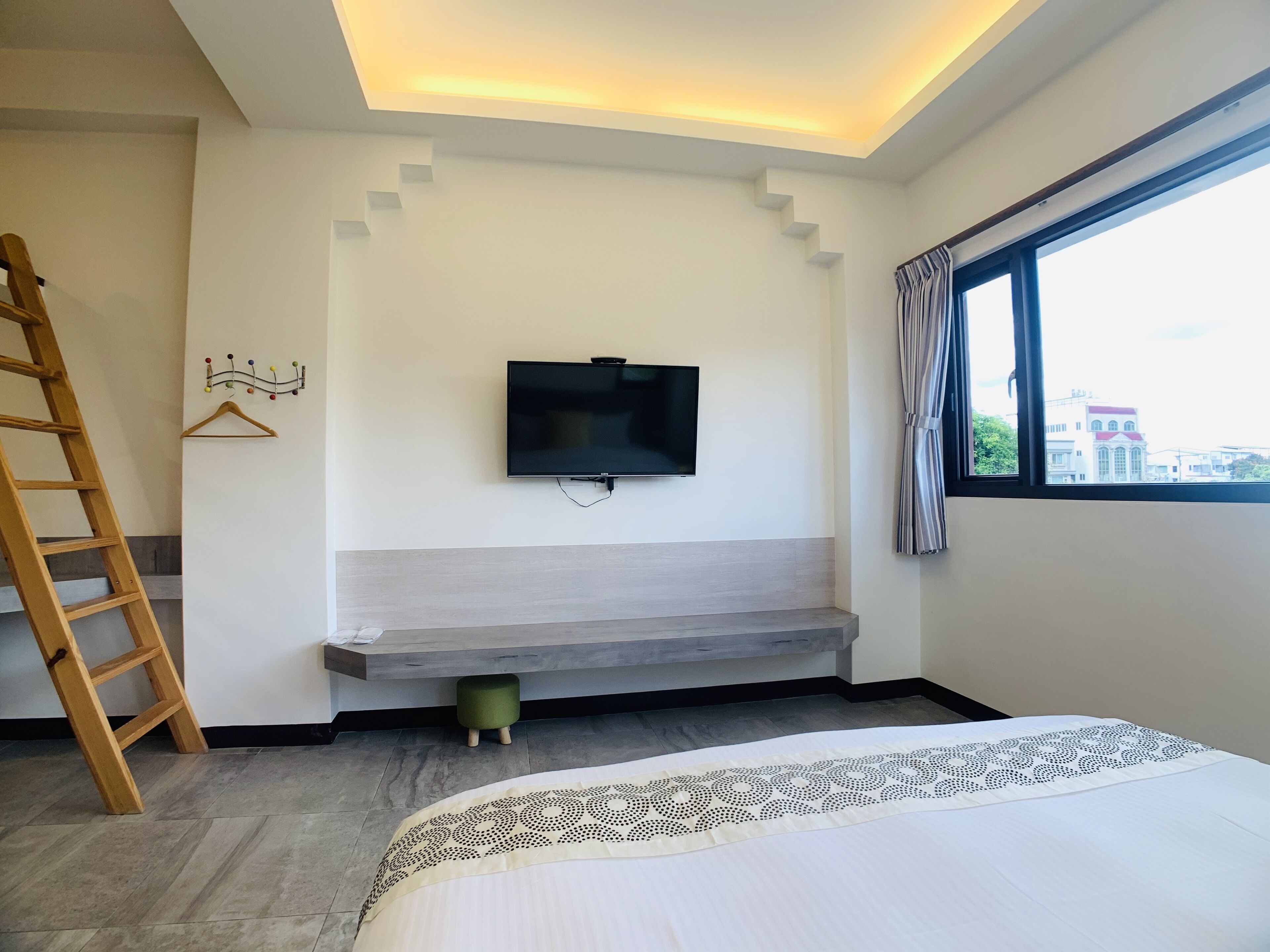 A bedroom with a bed, a TV mounted on the wall, a wooden ladder, a small stool, and a window with curtains.