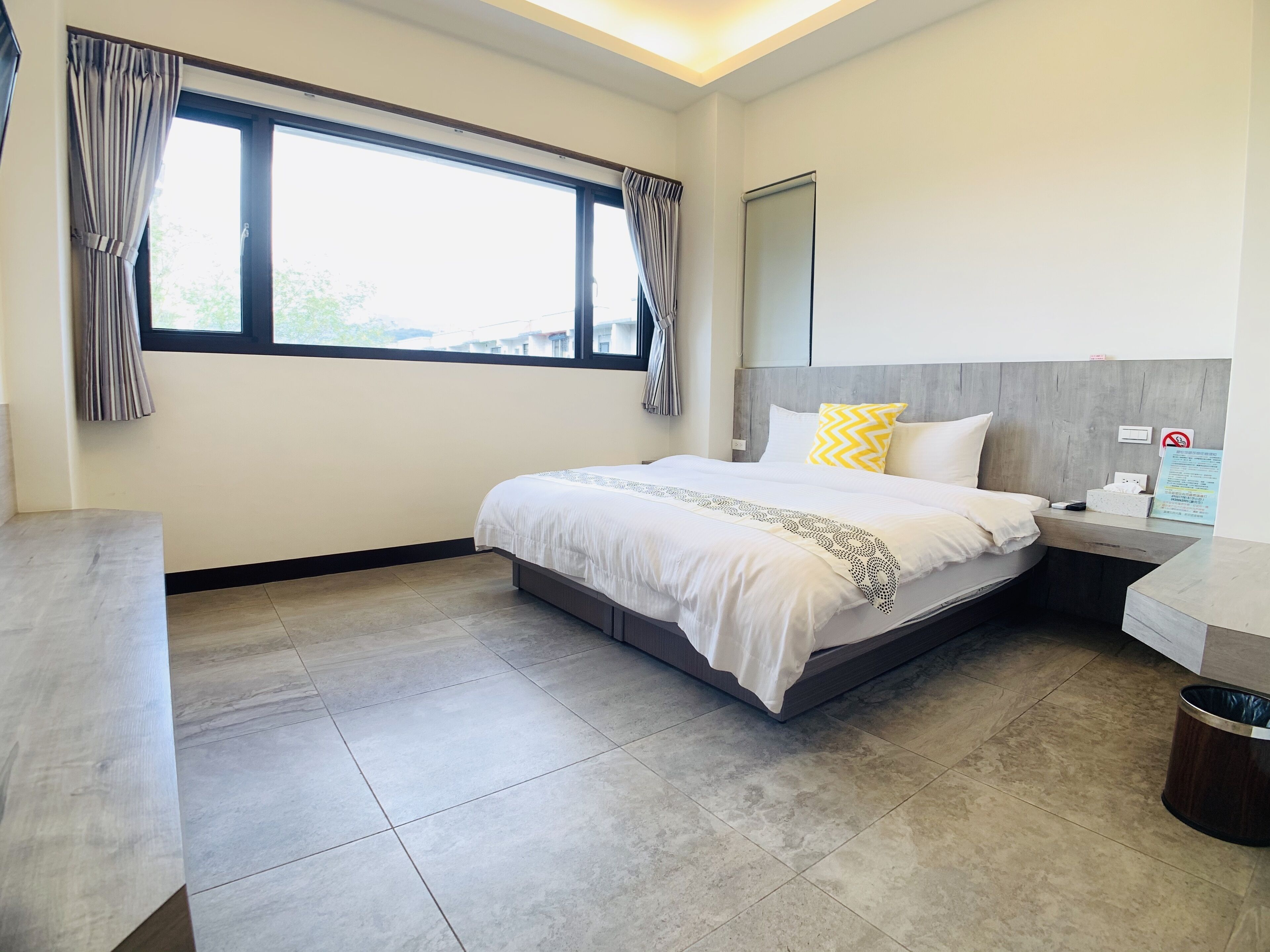 A modern hotel room with a large bed, a bedside table, a nightstand, a window with curtains, and a view of the outside.