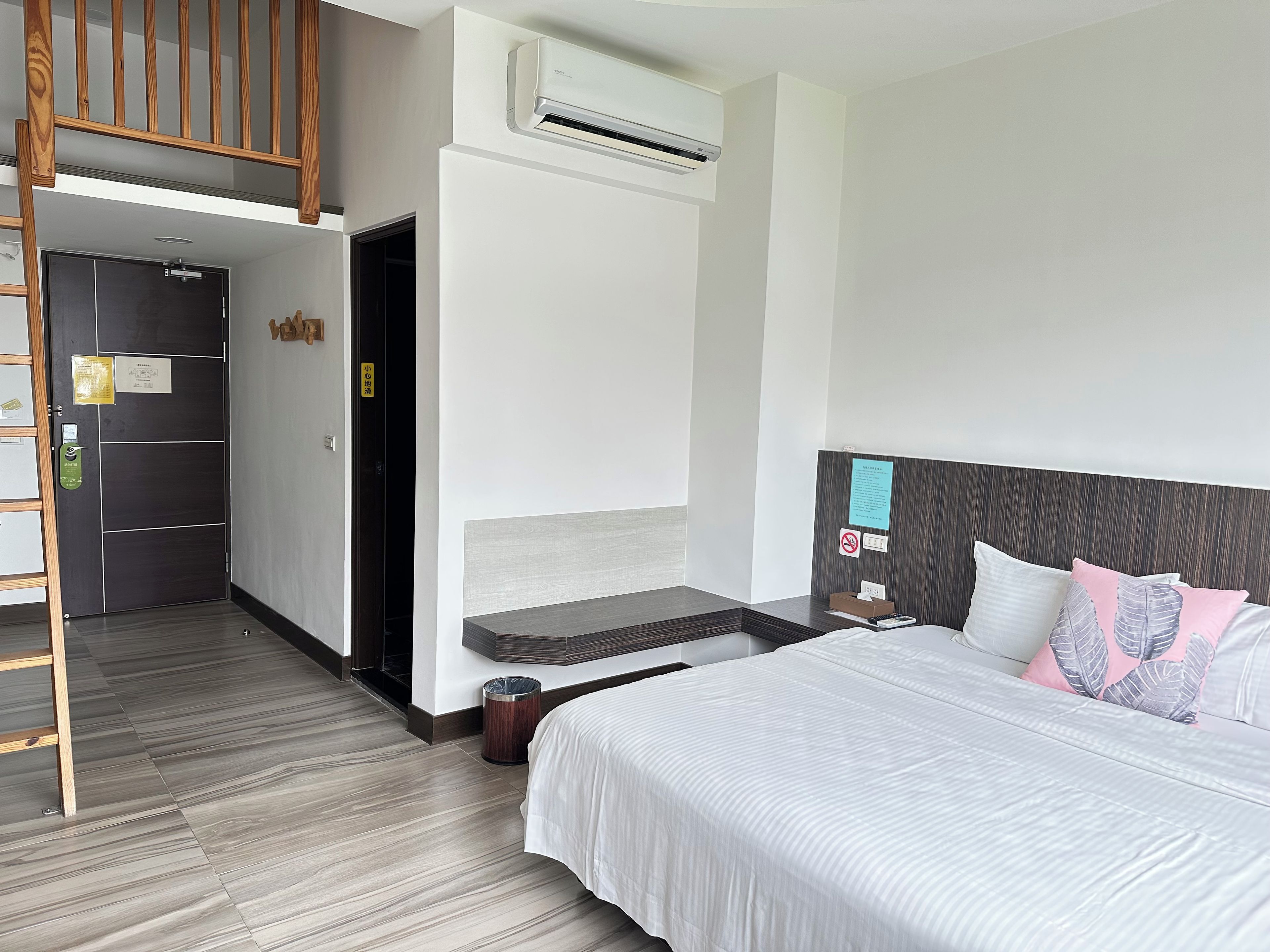 A hotel room with a bed, a desk, an air conditioning unit, a wooden staircase, and a door with a sign.
