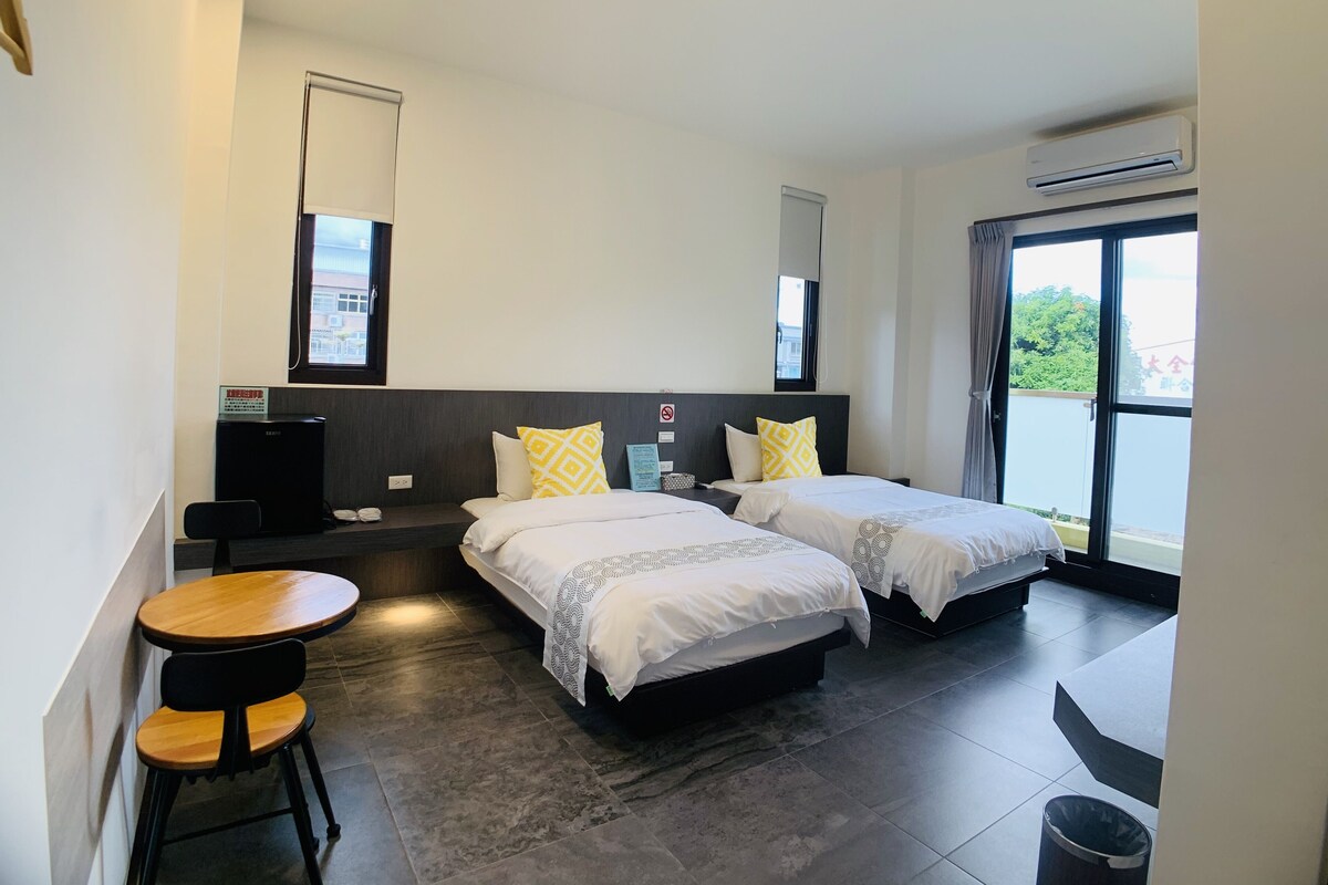 A hotel room with two beds, a small table with chairs, a desk, and a trash bin.
