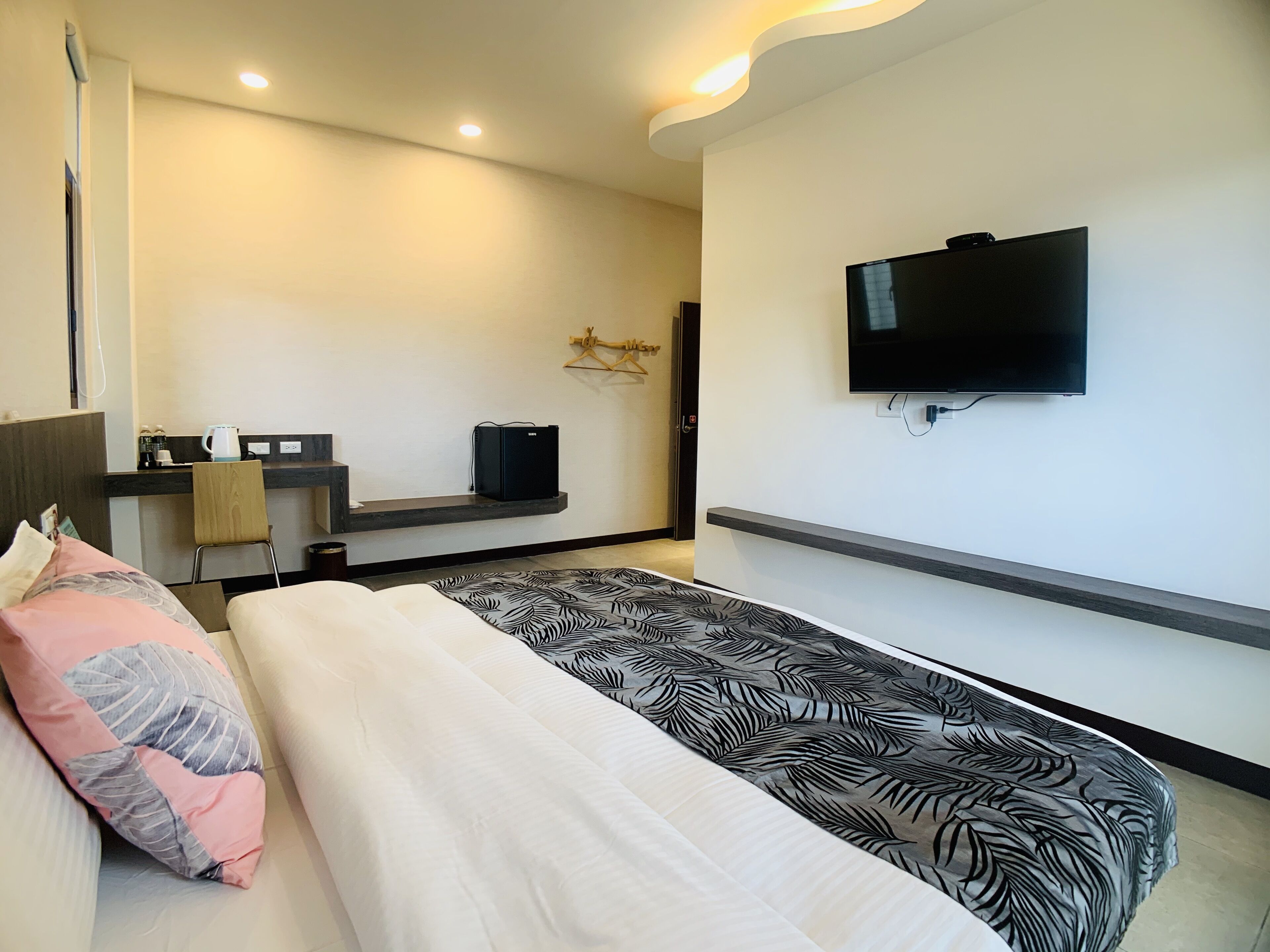 A modern hotel room with a bed, a TV mounted on the wall, a small desk with a kettle, and a chair.