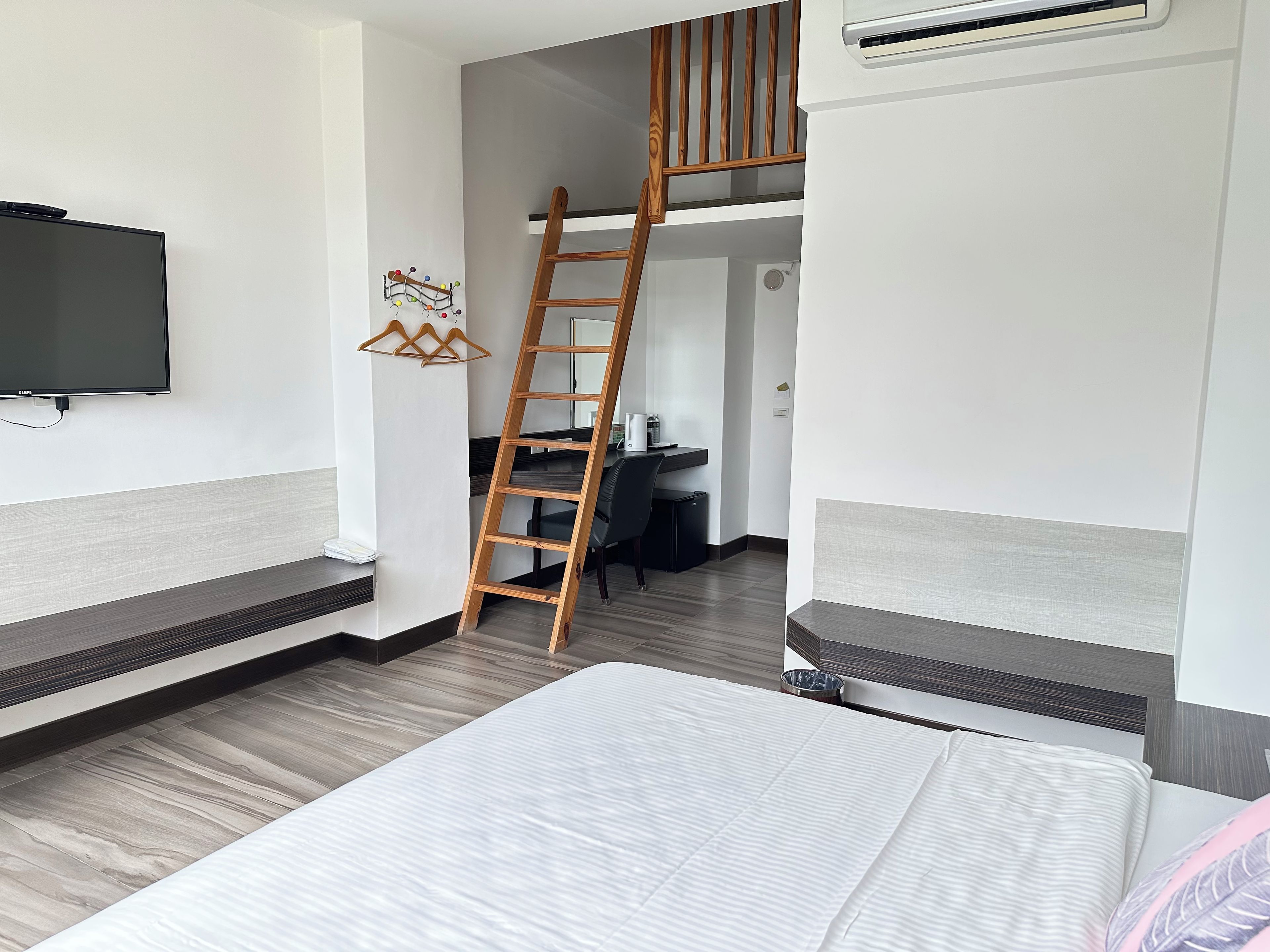 Habitación de hotel con cama, escritorio, silla, televisor montado en la pared y escalera de madera que lleva a un altillo.