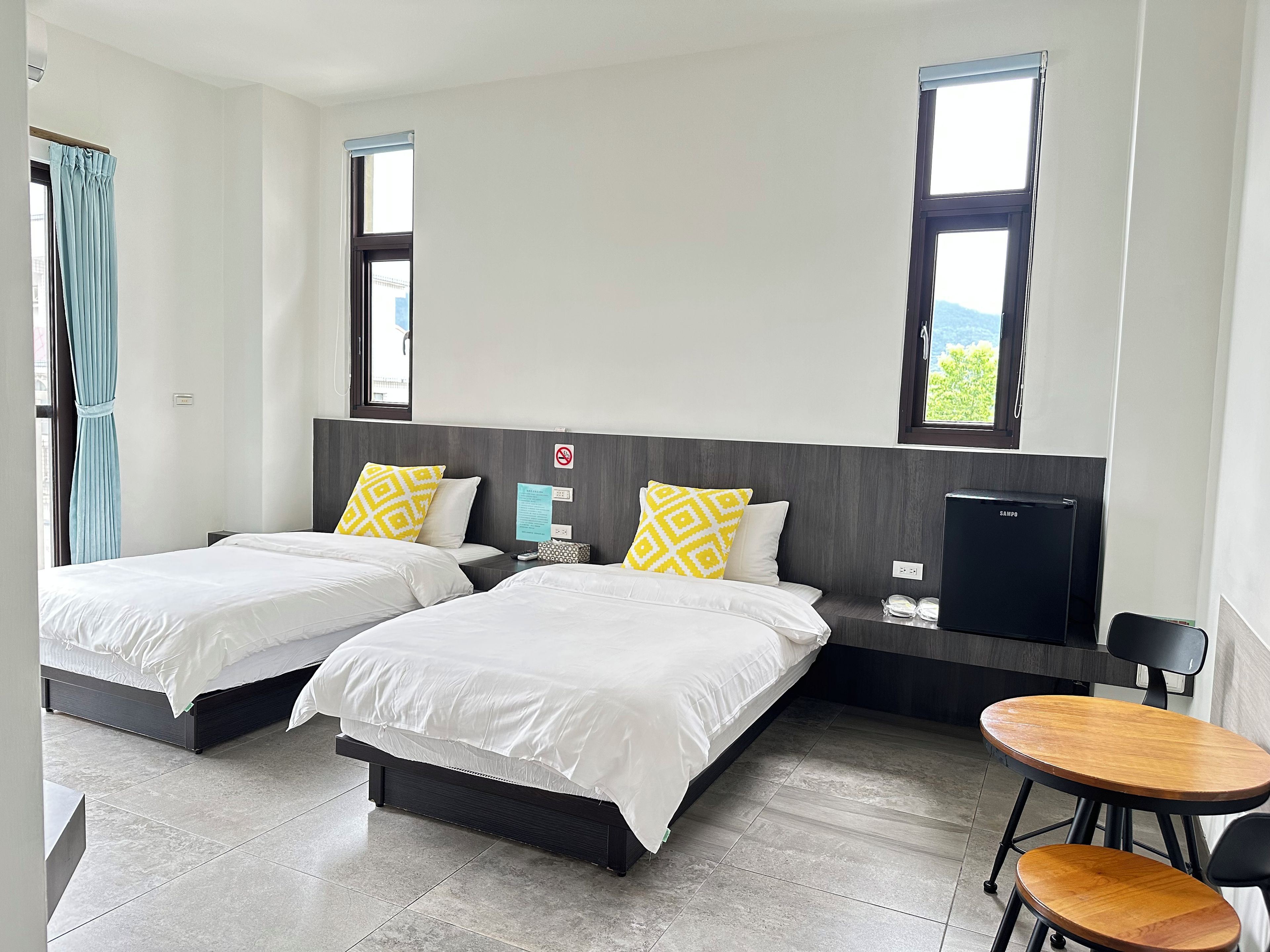 A hotel room with two beds, a desk, a chair, a minibar, and a view of the outdoors.