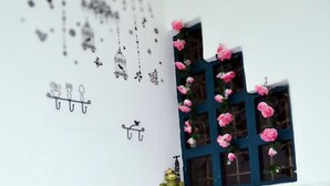 A room with a white wall decorated with wall art, a window with pink flowers, a pink chair with a decorative pillow, and a small green plant. - Manis-Manis Pink Lady Cottage (Malacca City)