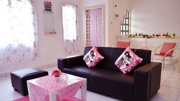 A living room with a dark sofa, two matching cushions, a small round table with a floral tablecloth, and a pink rug. - Manis-Manis Pink Lady Cottage (Malacca City)