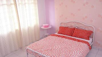 A bedroom with a white metal bed frame, a red polka dot bedspread, and matching pillows. There is a small round table with a pink object on it, and a white curtain with a wavy pattern.