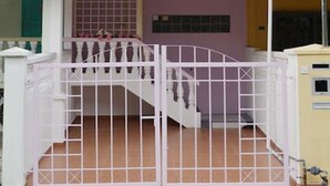 A two-story house with a white exterior, a balcony on the upper floor, and a gated entrance with a staircase leading to the main door. - Manis-Manis Pink Lady Cottage (Malacca City)