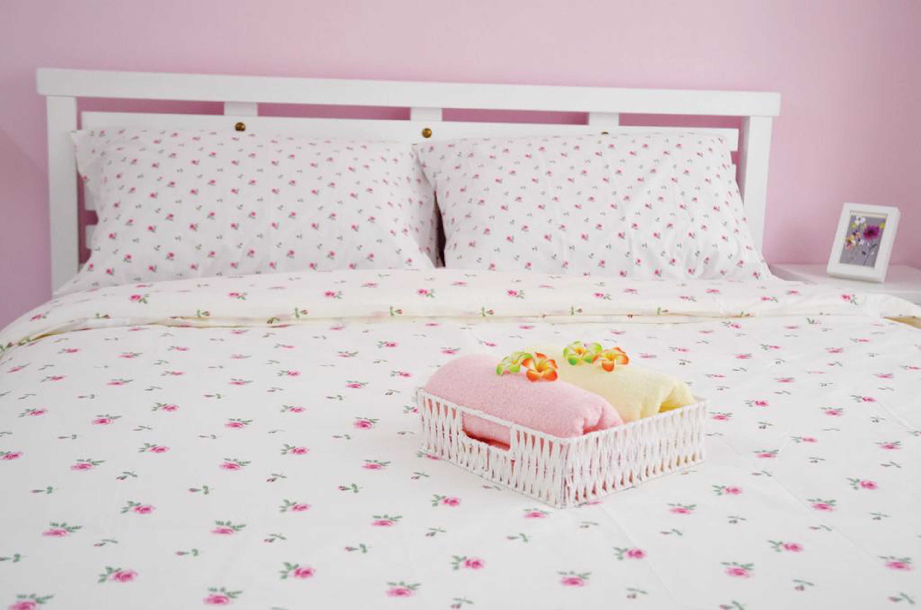 A bed with a floral patterned bedspread, white pillows with a small floral design, and a white bedside table with a picture frame.