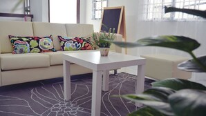 Una habitación con sofá color beige, almohadones de colores, una mesa blanca, una planta en maceta y un ventanal con vistas.