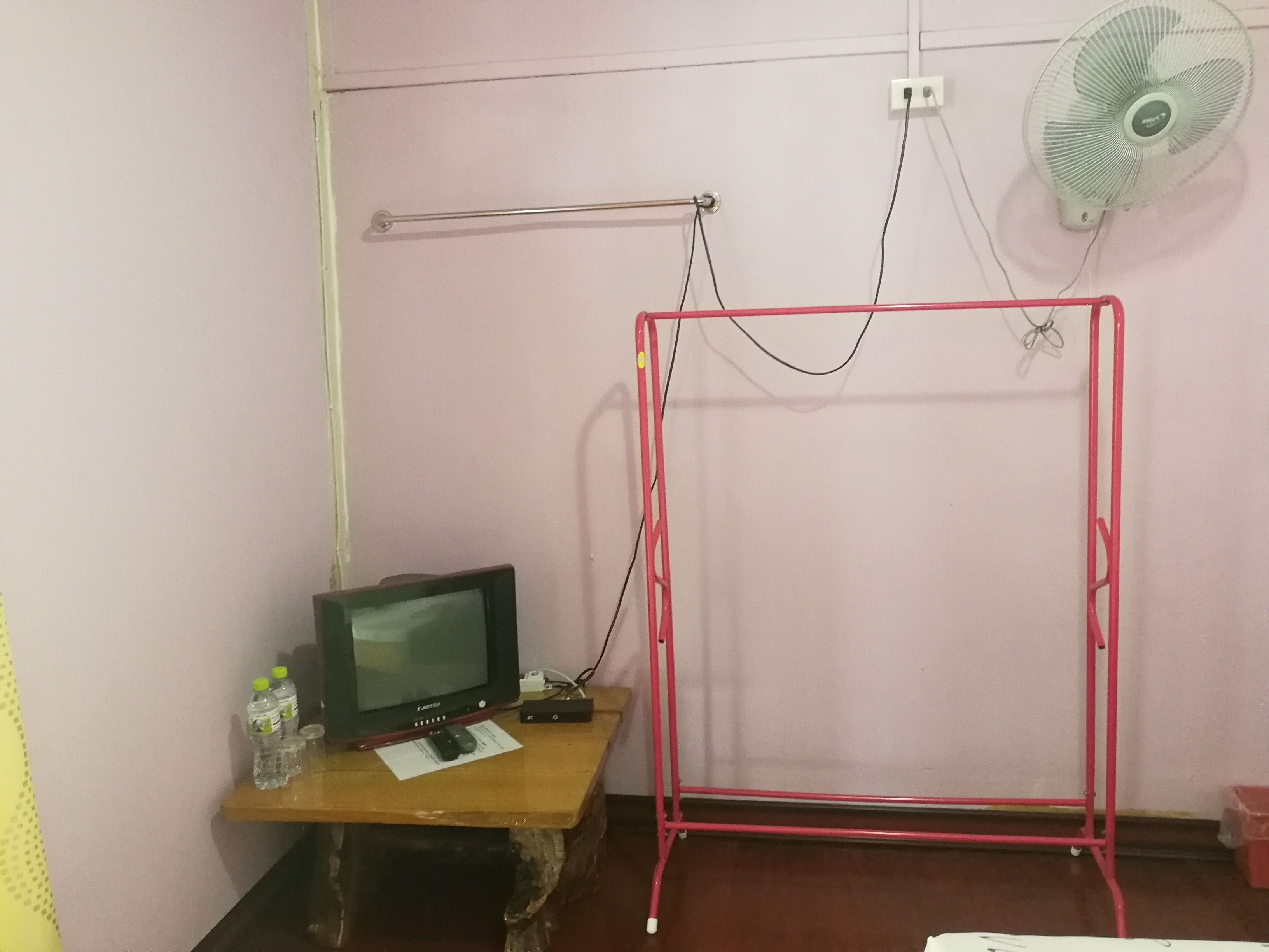 Ein Zimmer mit einem Holztisch, einem Röhrenmonitor, einem roten Metallständer, einer weißen Wand und einem Standventilator.