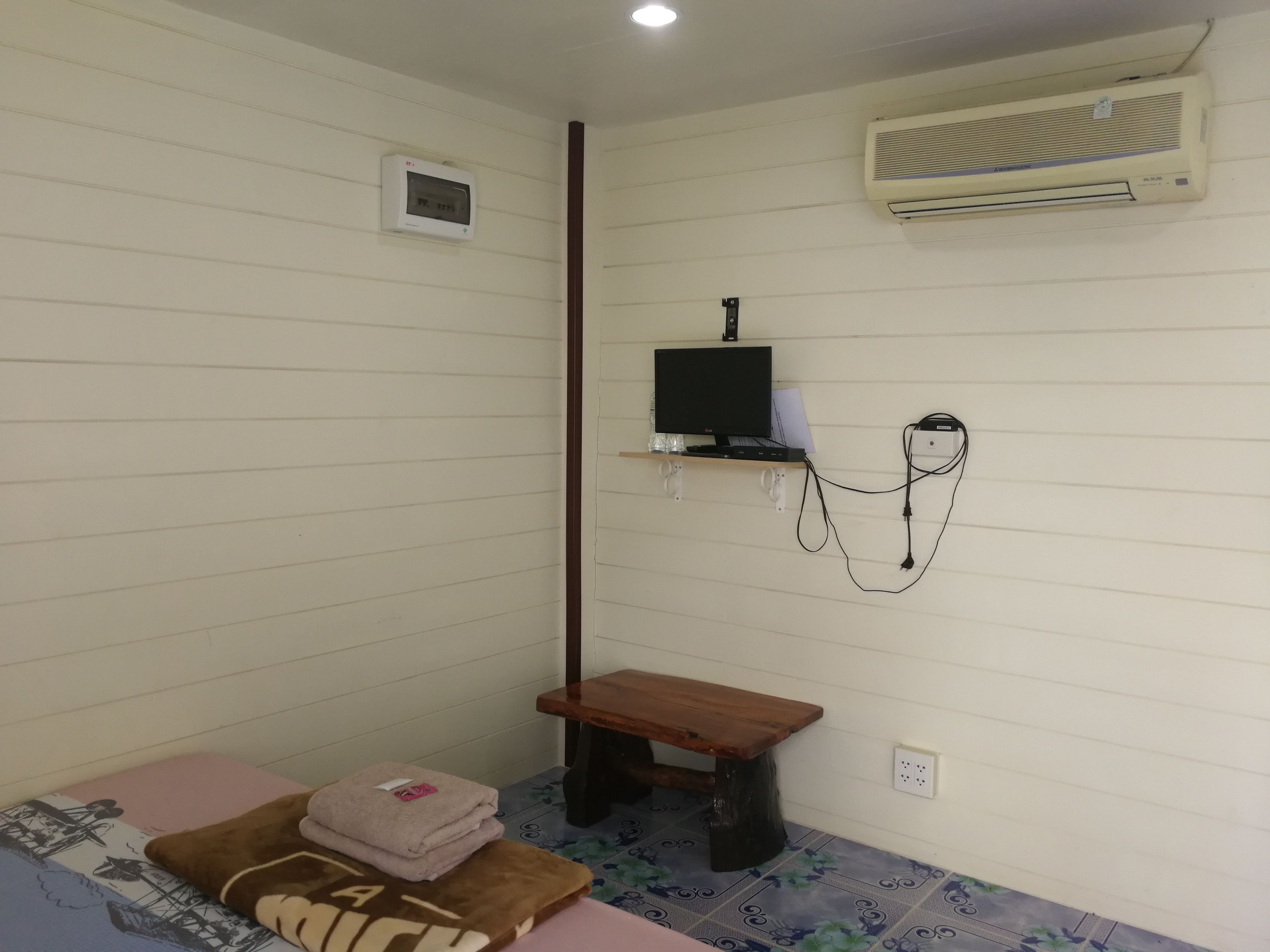 Une petite pièce simple, avec un lit, un banc, un téléviseur posé sur une étagère, un climatiseur et un thermostat.
