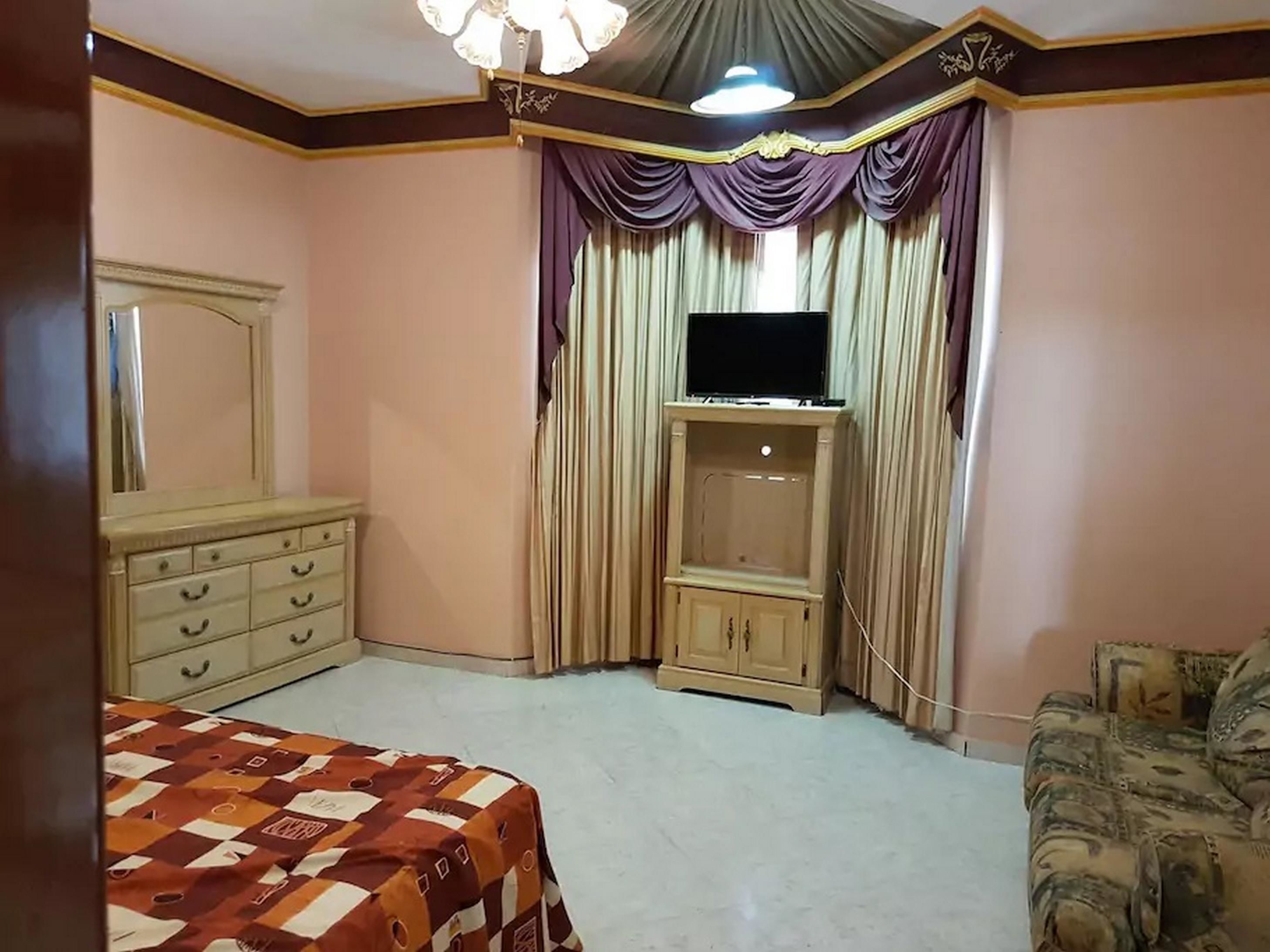 A bedroom with a bed, a dresser, a TV on a stand, and a patterned rug.