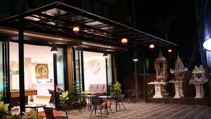 Un espacio al aire libre con mesas y sillas, una sección cubierta con un muro de vidrio y un santuario decorativo.