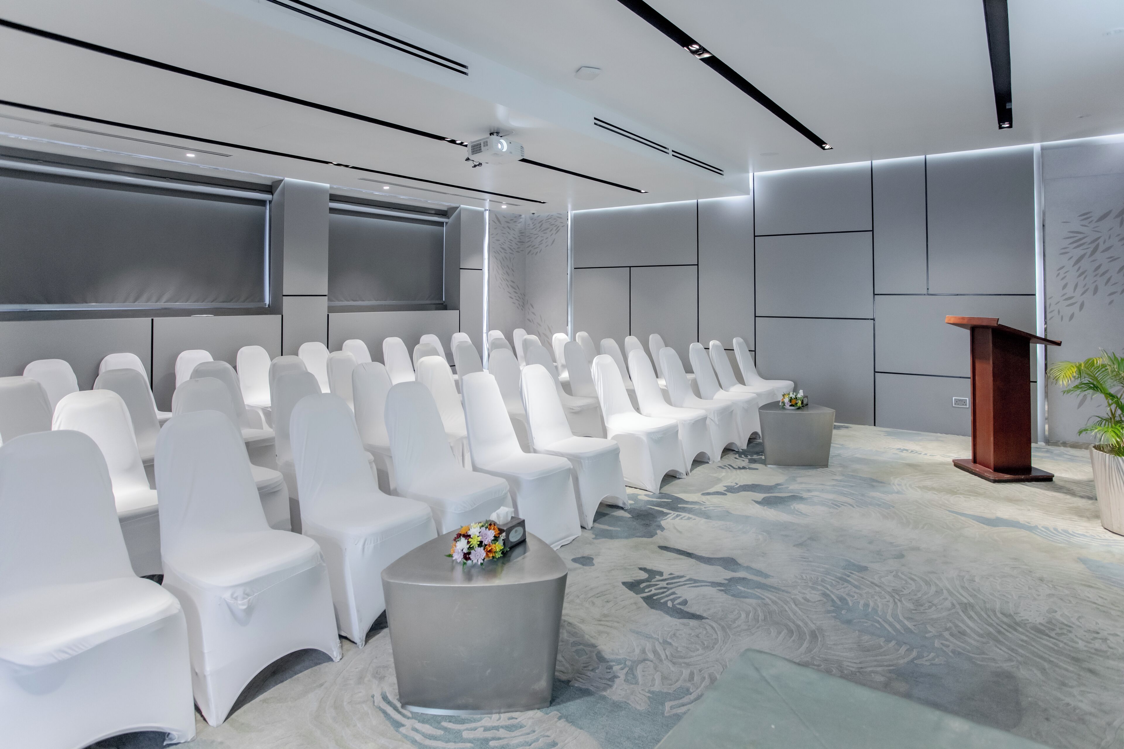 Une salle de conférence moderne, dotée de chaises blanches, d’un pupitre et d’un écran de projection.