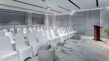 Une salle de conférence moderne, dotée de chaises blanches, d’un pupitre et d’un écran de projection.