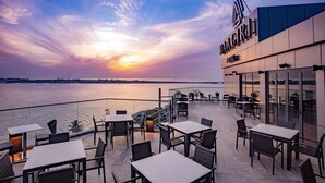Pérgola de hotel frente al agua con mesas y sillas, barandilla de vidrio y vista del atardecer sobre el agua.