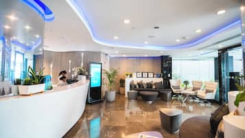 Eine moderne Hotelhalle mit einer Rezeption, einer Sitzecke und einer großen digitalen Anzeige.