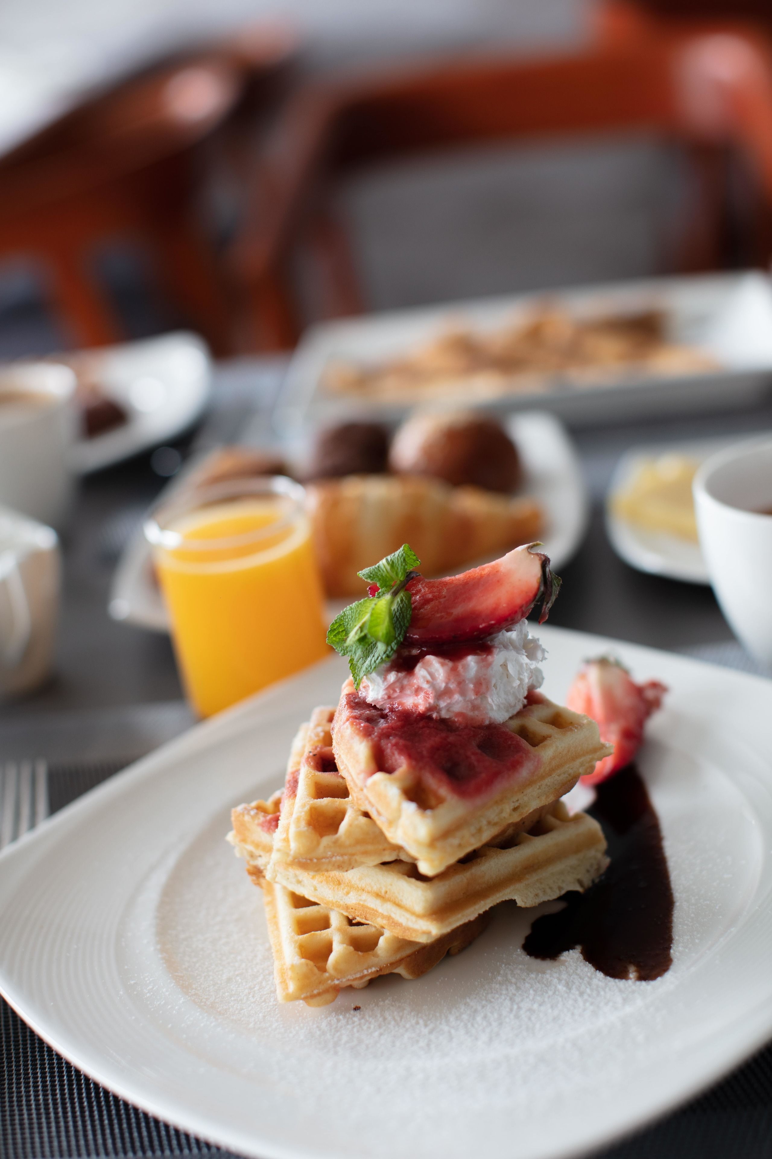 Une table dressée pour le petit-déjeuner avec des gaufres garnies de fraises et de crème fouettée, un verre de jus d’orange et d’autres aliments typiques du petit-déjeuner.