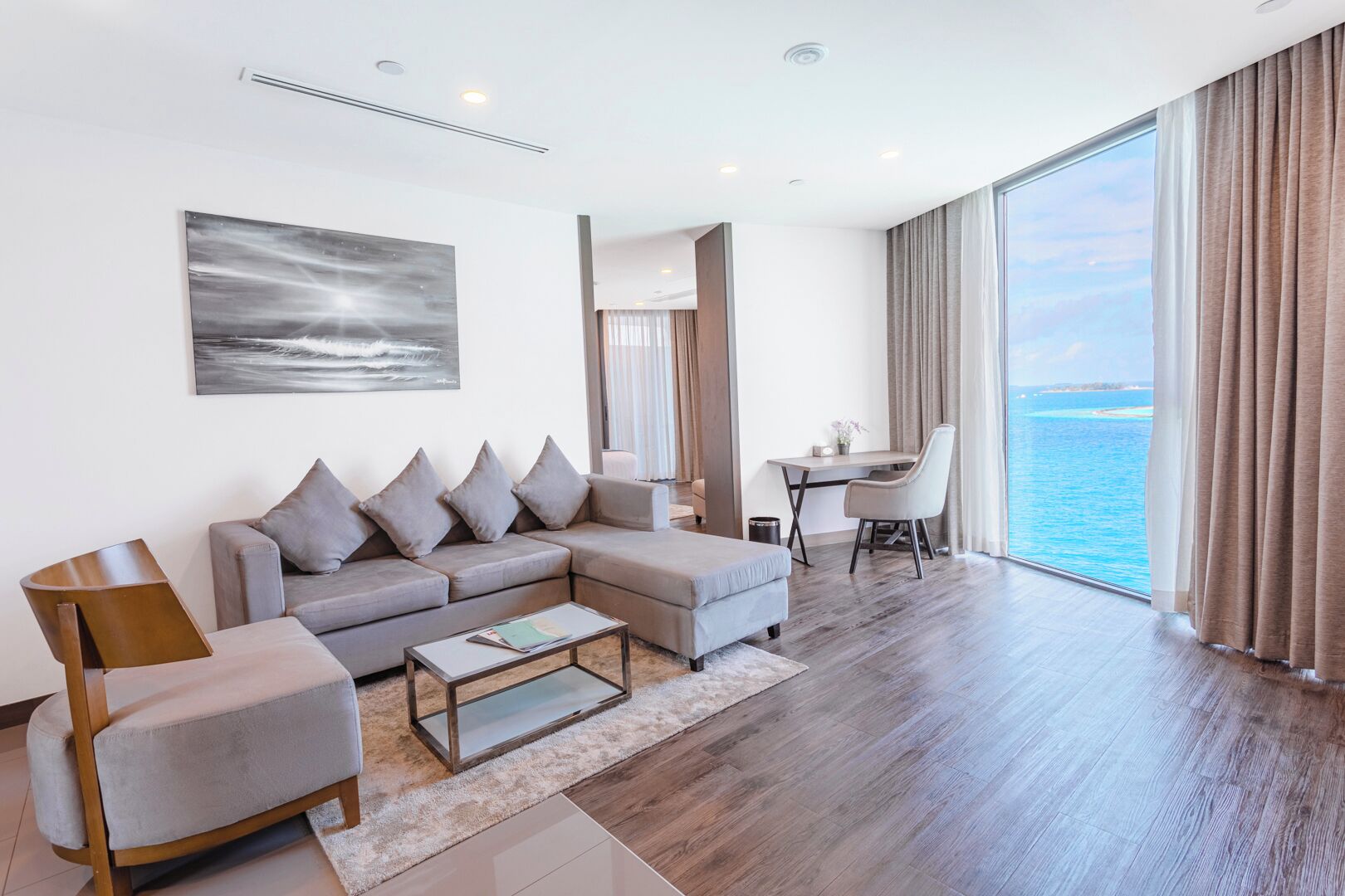 A modern living room with a sofa, armchair, and coffee table, a desk with a chair, and a large window overlooking the sea.