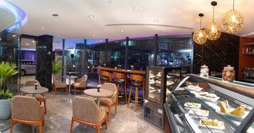 Un café moderno con mesas y sillas redondas, un expositor de vidrio con pasteles, y una zona de estar con vistas a la calle.