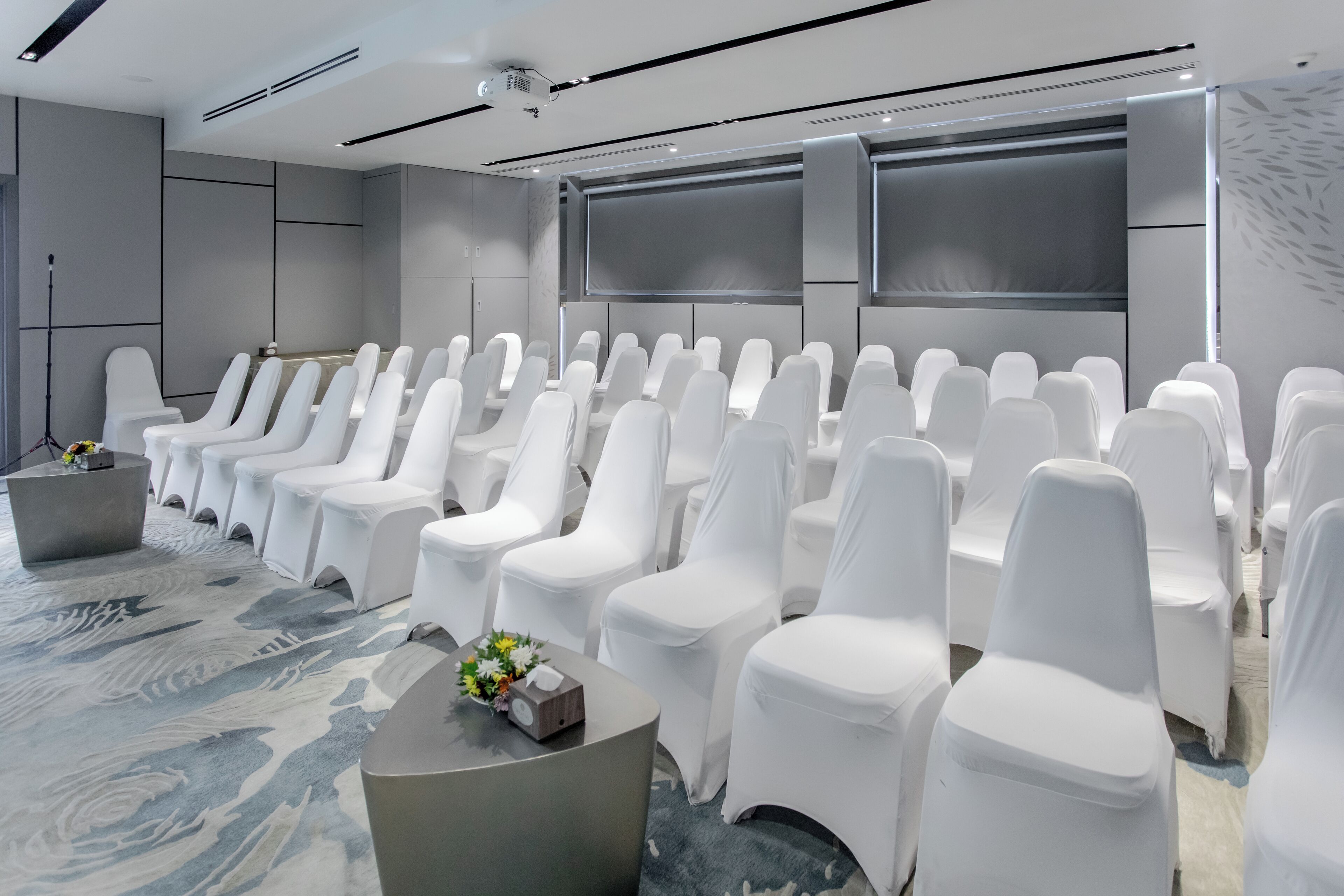 Une salle de conférence avec des rangées de chaises blanches, un écran de projection et un tapis à motifs de marbre.