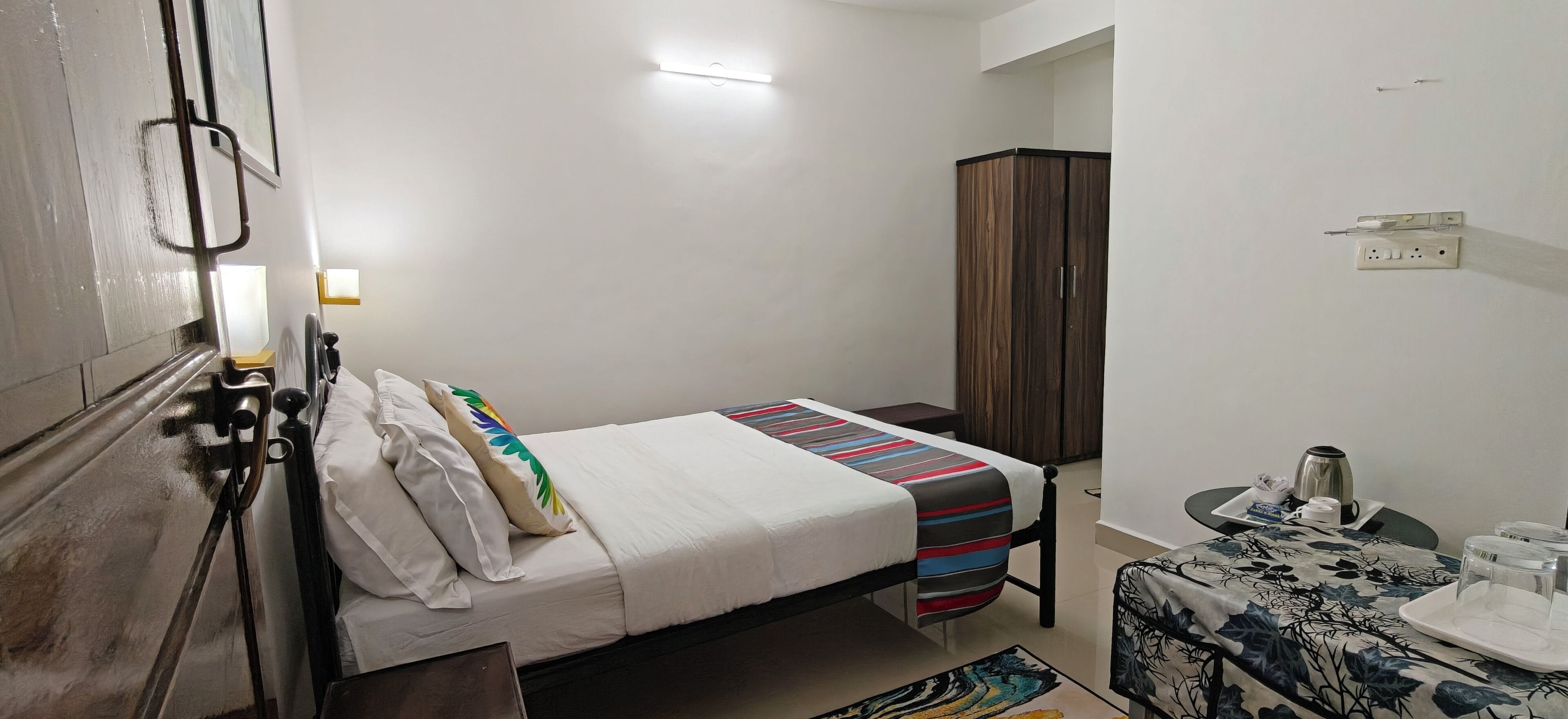 Ein Einzelbett mit gestreifter Decke, ein kleiner Nachttisch mit Wasserkocher, ein Schrank und eine Wandleuchte.