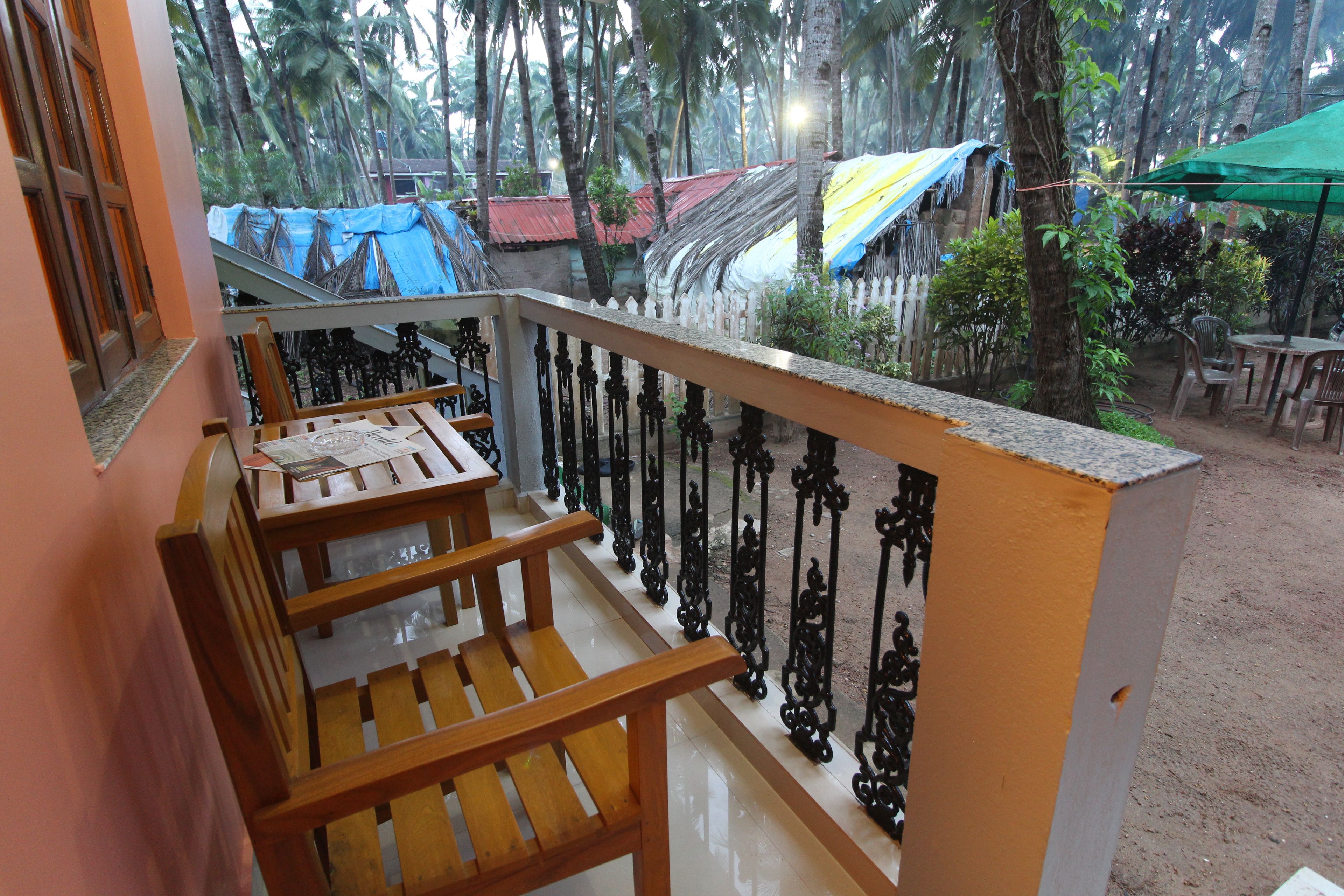 A balcony with wooden chairs and a table, overlooking a garden with palm trees and covered structures.