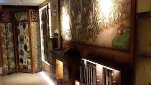 A hallway with a decorative wall featuring a mural of people, a counter with a green lamp, and a patterned wall on the left. - IND Hotel (Kowloon)