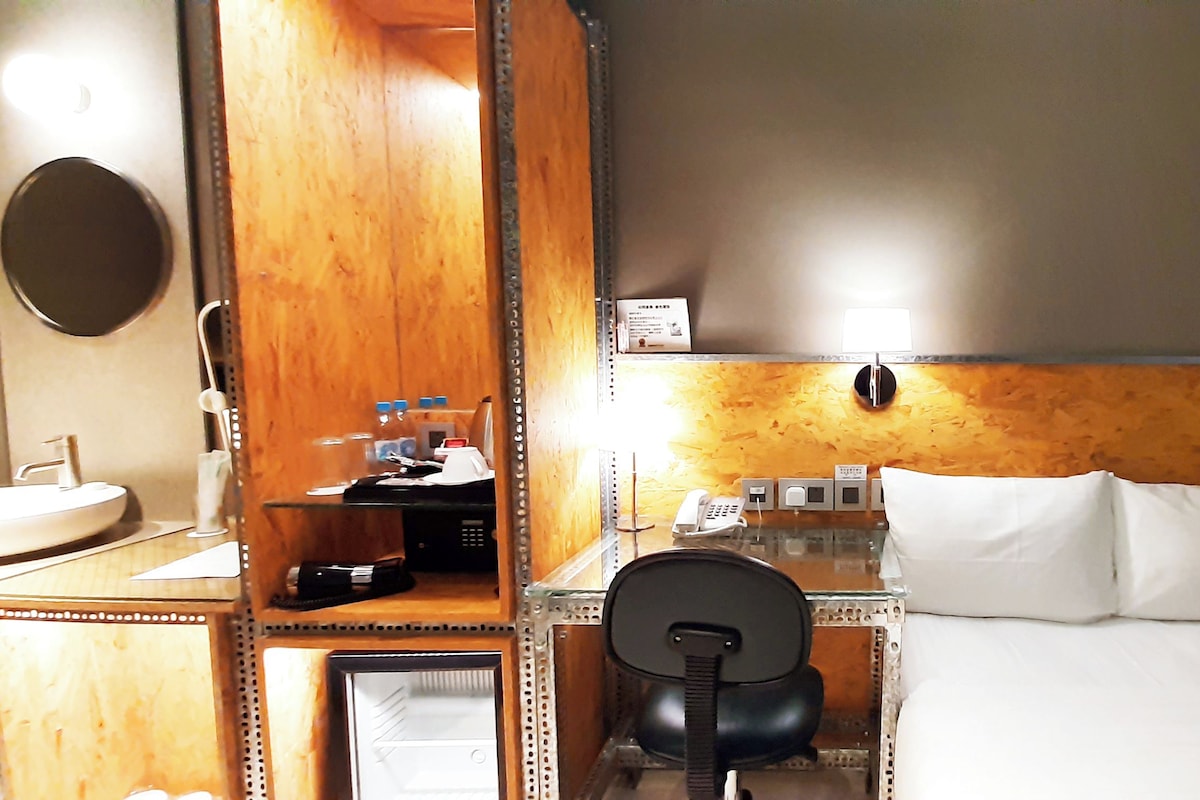 Une chambre d’hôtel avec une tête de lit en bois, un bureau avec un téléphone, une chaise, un lit avec du linge de lit blanc et un miroir rond accroché au mur.