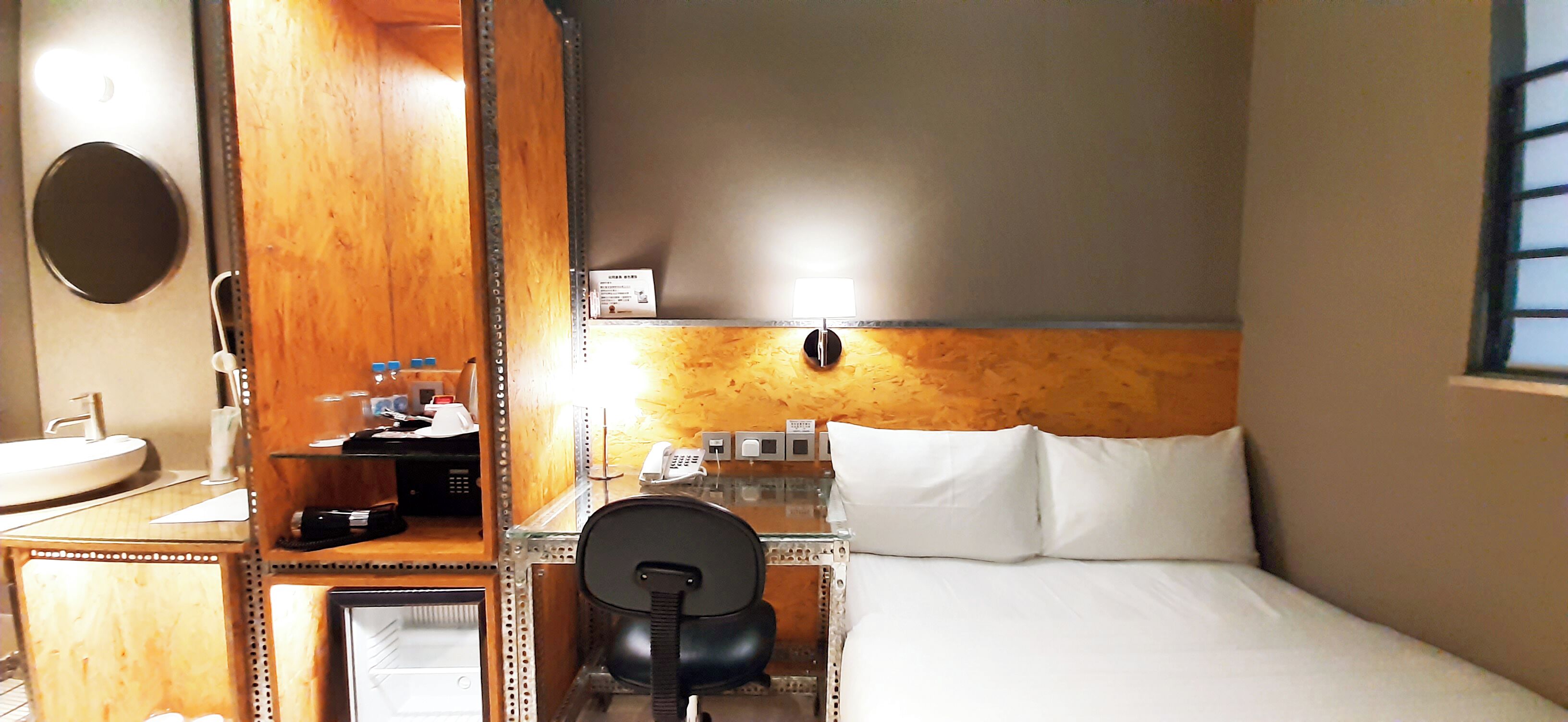 Een hotelkamer met een houten hoofdbord, een bureau met telefoon, een stoel, een bed met witte lakens en een ronde spiegel aan de muur.