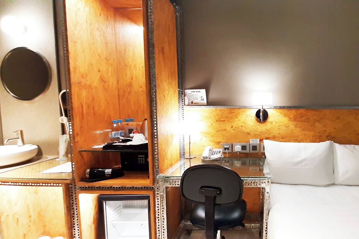 A hotel room with a wooden headboard, a desk with a telephone, a chair, a bed with white linens, and a round mirror on the wall.