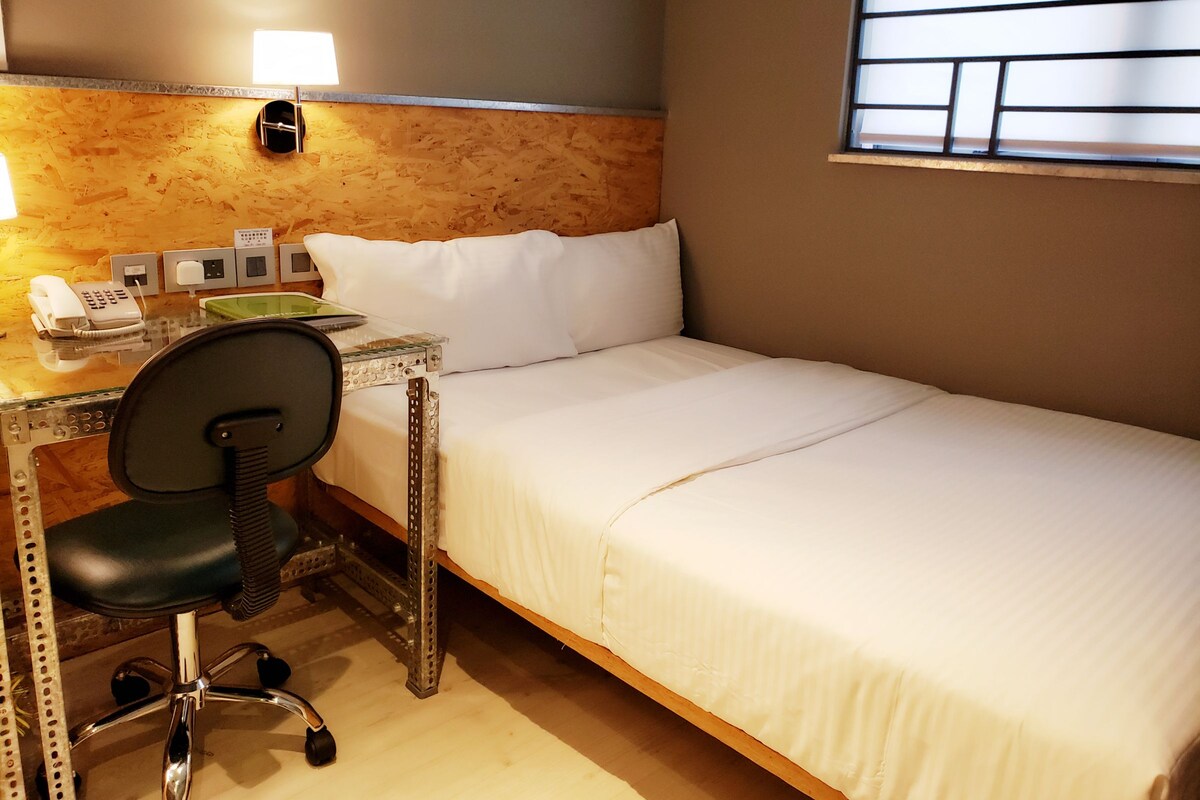 A hotel room with a bed, a desk with a telephone, a chair, and a window with blinds.