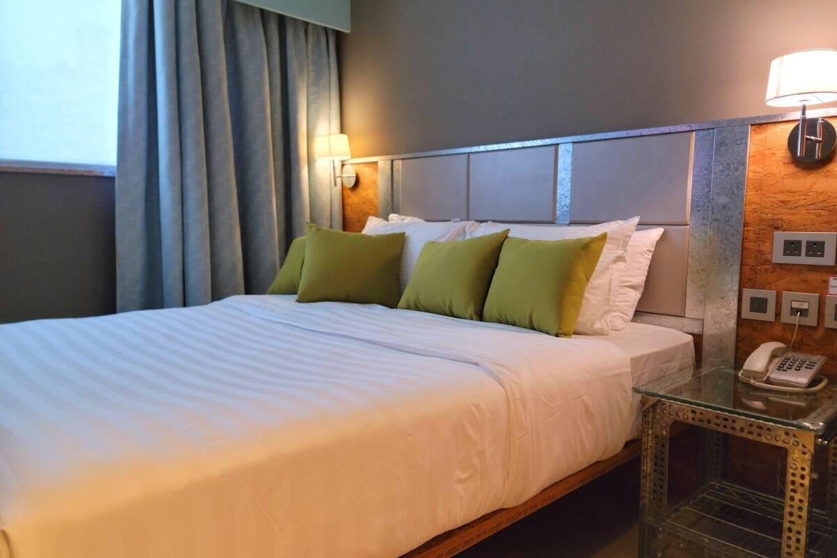 A hotel room with a bed, green pillows, a bedside lamp, a telephone, and a wall-mounted light switch.