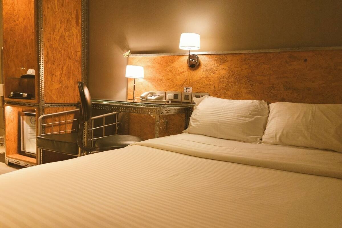 A hotel room with a bed, a desk with a telephone, a chair, and a wall-mounted lamp.