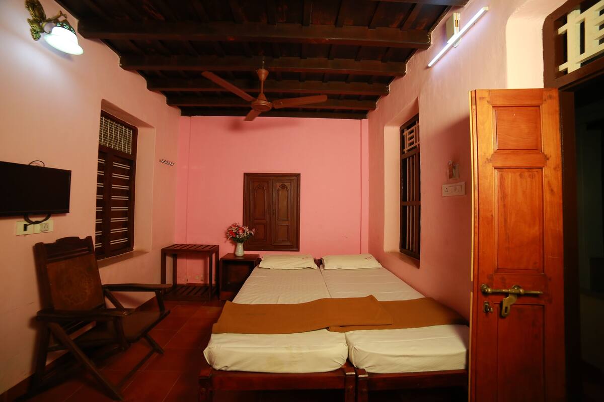 A room with two beds, a wooden door, a ceiling fan, a TV mounted on the wall, and a window with shutters.