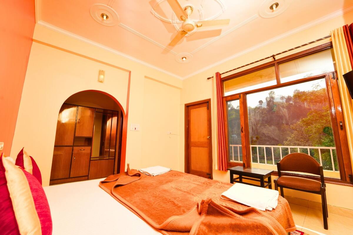 A hotel room with a bed, a desk, a chair, a window with a view of trees, and a ceiling fan.
