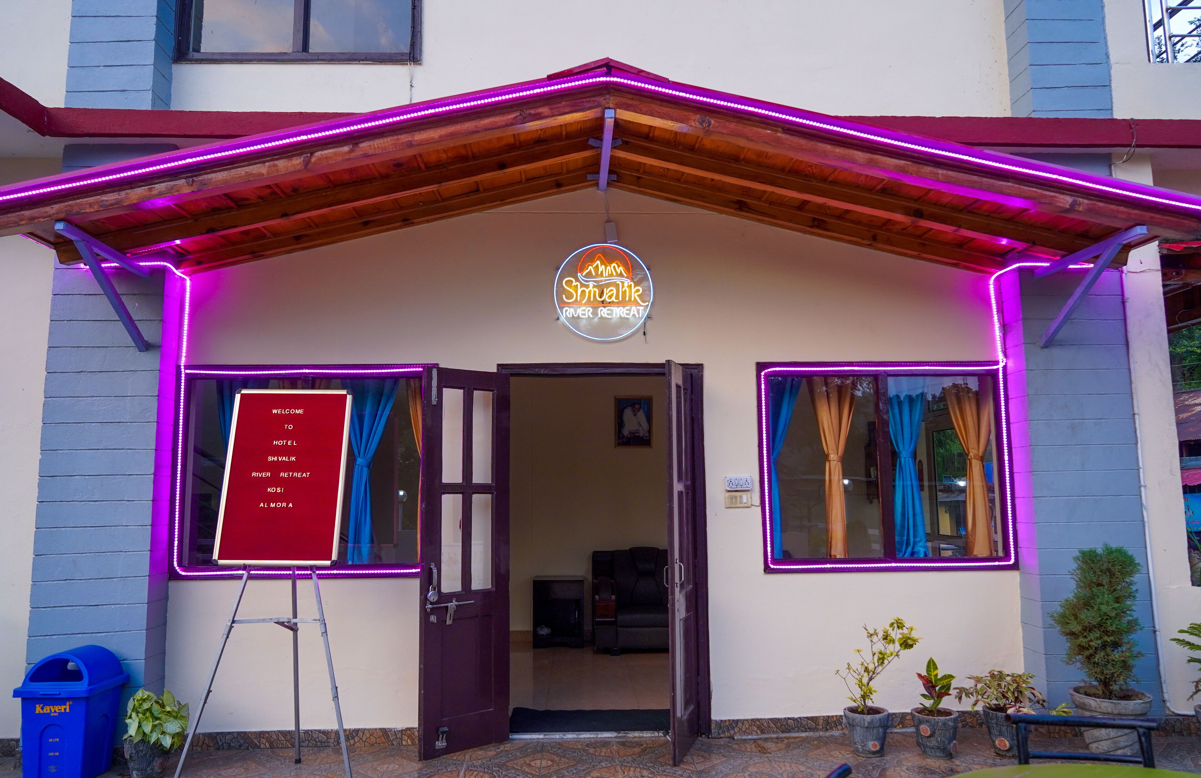 Ingresso di un hotel con un neon, un cartello di benvenuto e piante in vaso.