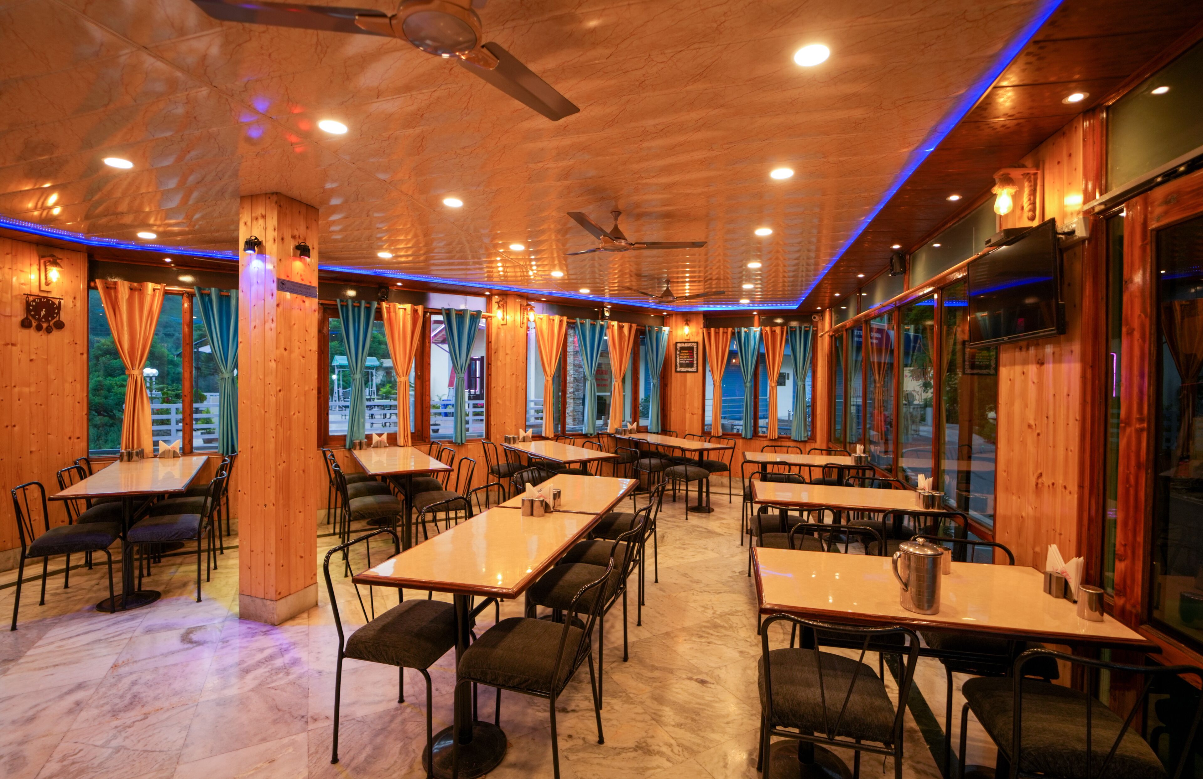 Un ristorante con arredamento in legno, diversi tavoli e sedie, ventilatori a soffitto e ampie finestre con tende.