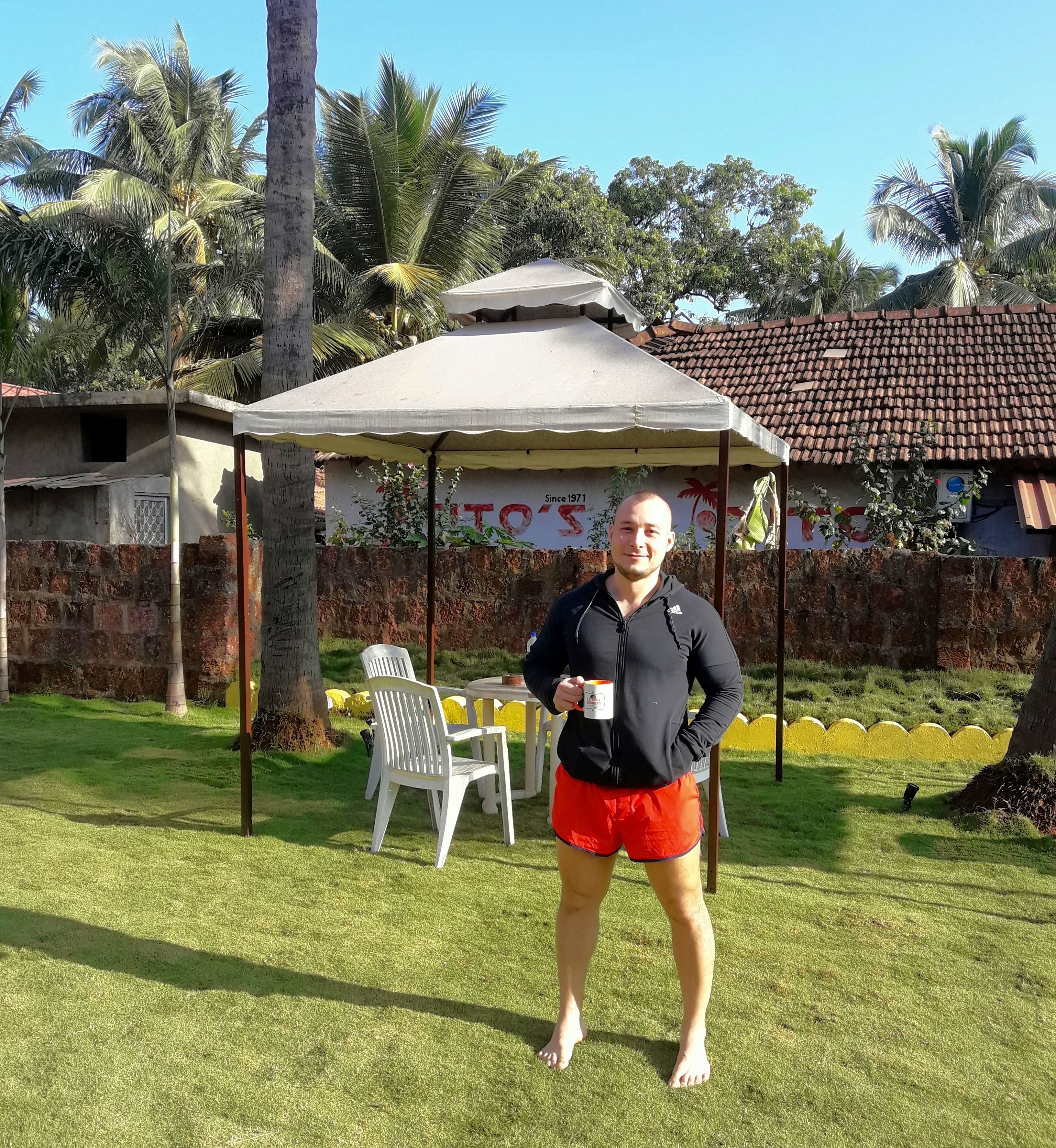 A person standing in a grassy area with a white gazebo, a table with chairs, and a building with a tiled roof in the background.