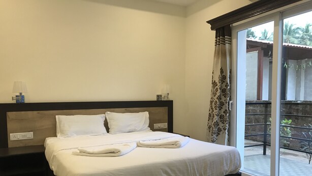 A neatly made bed with white linens and towels, a bedside table with a lamp, and a view of a balcony with plants and a building in the background. - La Vega Resort (Morjim)