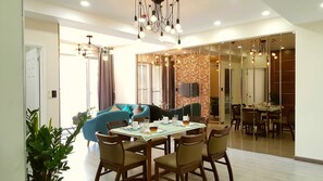 A dining area with a round table, chairs, and a chandelier. There is a sofa and a TV in the background.