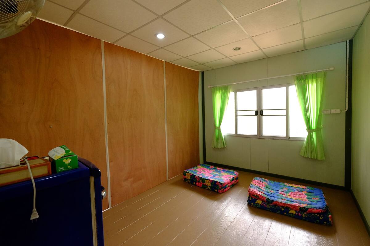 Ein Zimmer mit Holzboden, einem blauen Schrank, einem Ventilator und zwei bunten Matratzen.