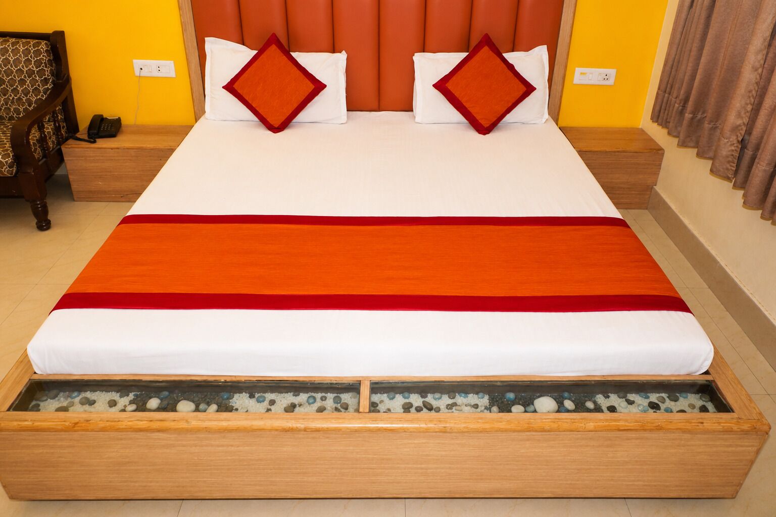 Una cama con colchón blanco, colcha naranja y roja, y dos almohadas con bordes naranjas.