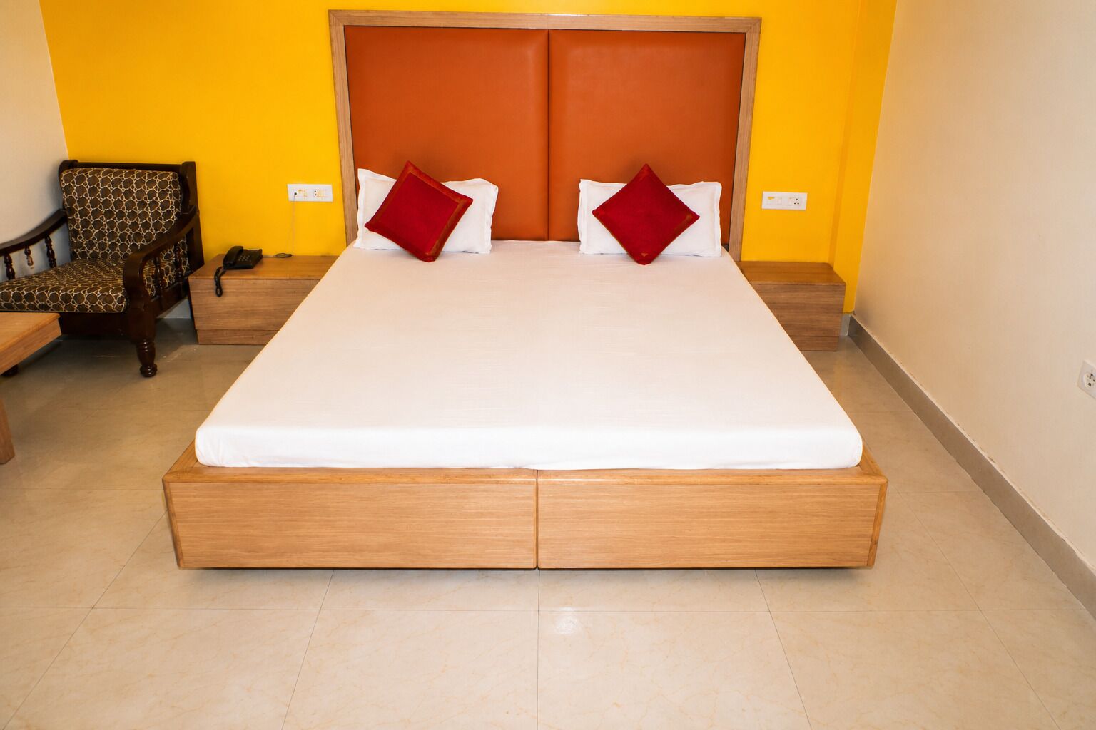 A bed with a wooden frame and two pillows, a chair with a patterned cushion, and a bedside table with a phone.