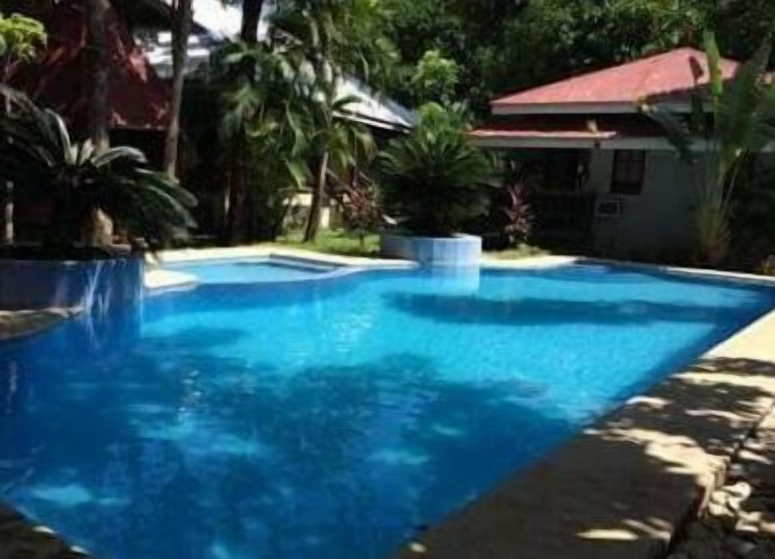 Una piscina azul cristalina rodeada de abundante vegetación y una casa con techo rojo.