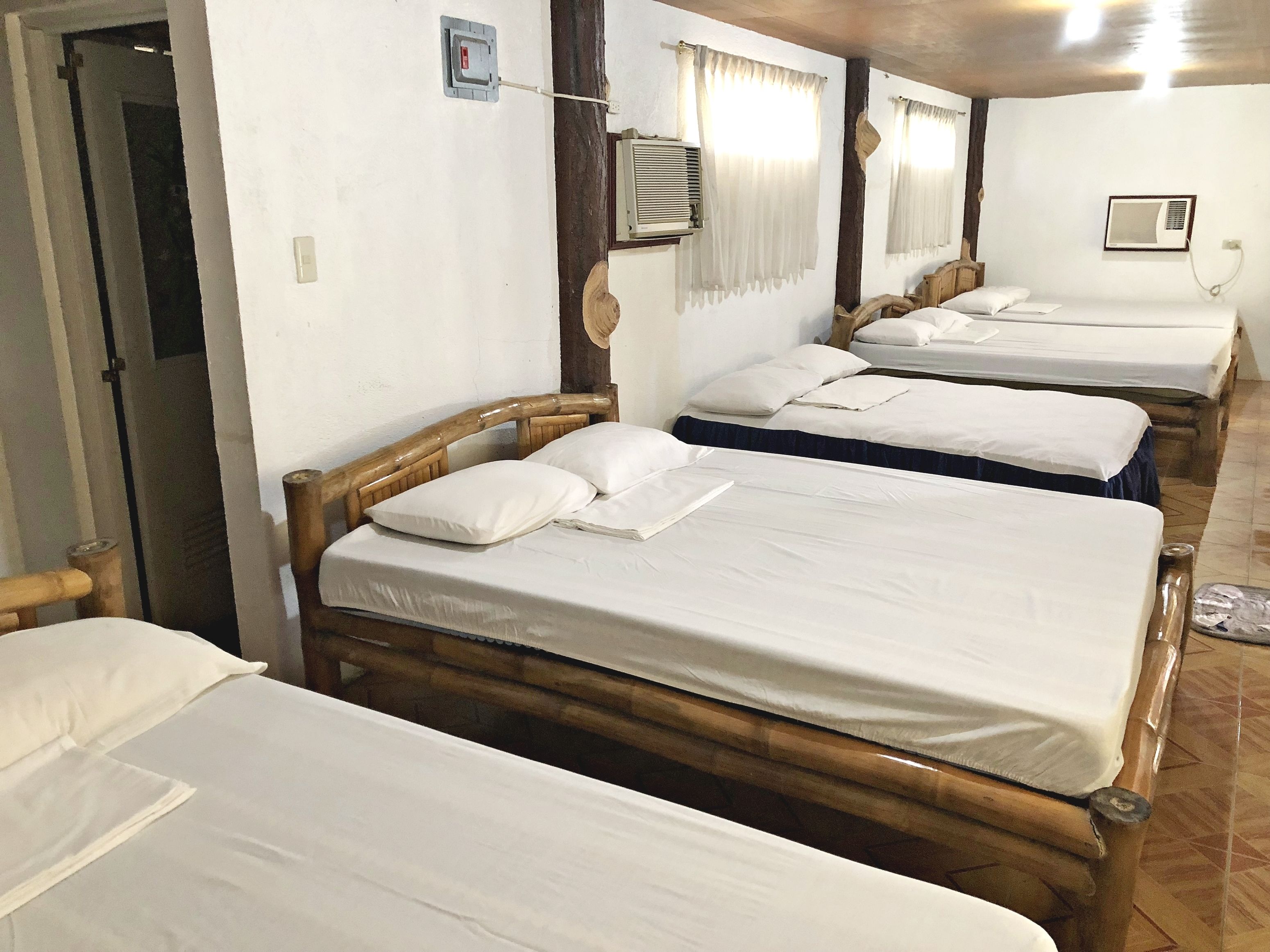 A room with four wooden beds, each with white linens and pillows. There are air conditioning units on the walls, and a door leading to another area.