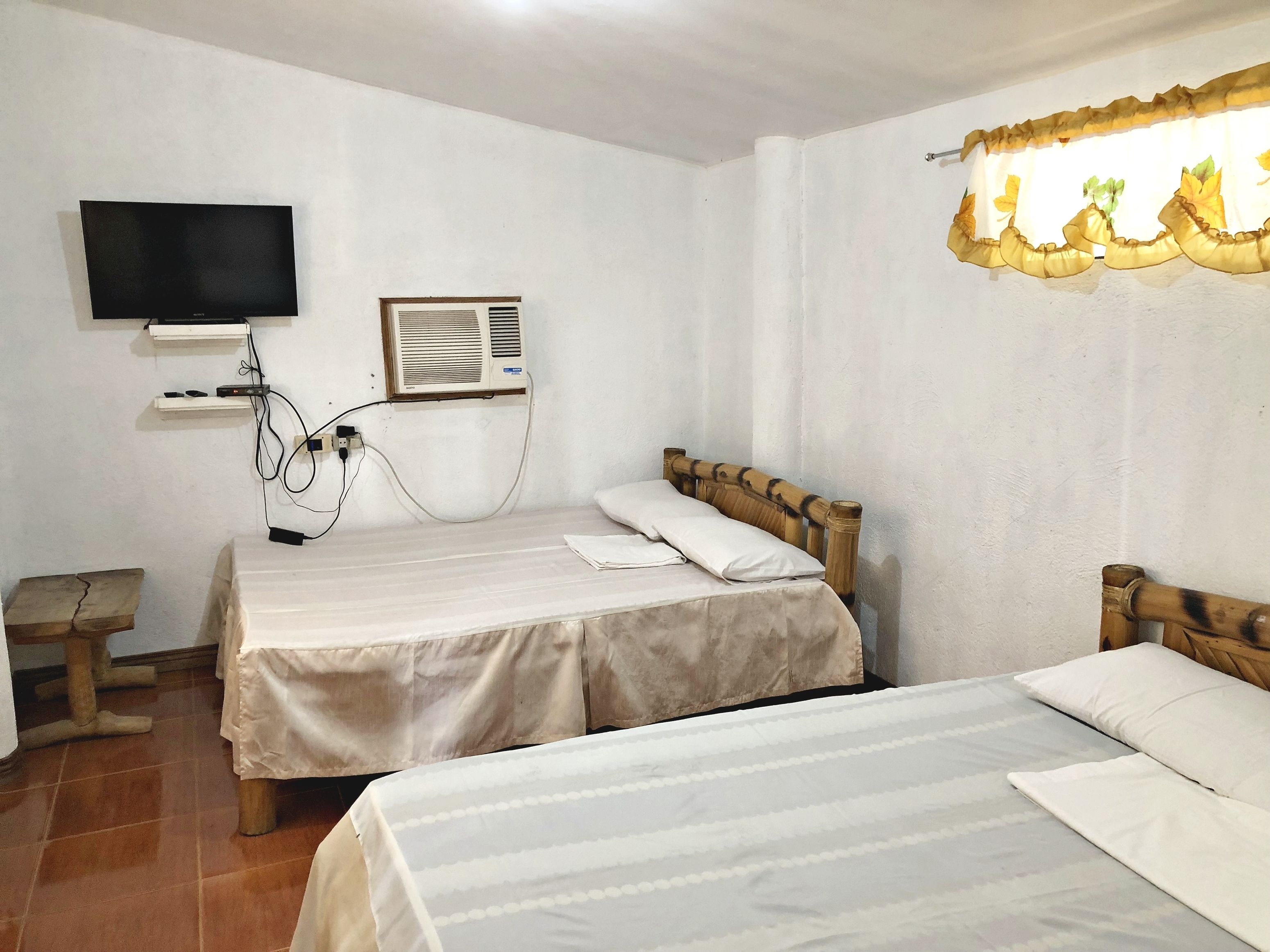 A room with two beds, a television mounted on the wall, and a window with a curtain.